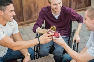https://unsplash.com/@elevatebeer A group of 3 happy looking men are clinking their glasses together and smiling. One of the men is seated in a wheelchair.