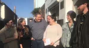 A group of 6 people are stood outside a building on a sunny day. They are all dressed casually and are smiling at each other. The woman in the centre is holding a piece of paper.