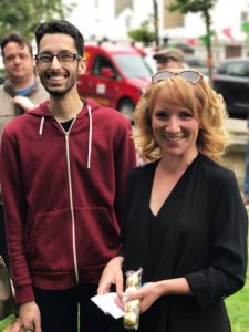 A young man and woman are standing outside on grass, you can see the street behind them and it is sunny. They are both smiling widely and looking at the camera.