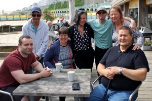 A group of 7 people are seated and standing around a table, smiling at the camera with their arms around each other. The table is outside on a decked area, with Brighton seafront behind them.