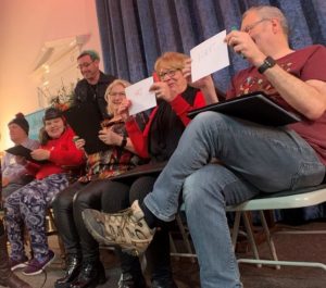 Five people are sat on chairs holding up pieces of cardboard, they are all smiling and laughing. One woman is wearing a soft helmet and a smiling man is stood behind her, supporting her. The people appear to be on a stage with a blue curtain behind them. Everyone looks like they’re having a good time.