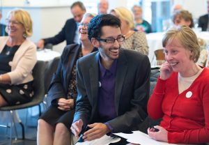 A young man seated at a busy event is smiling at a young woman sat next to him. They both look very happy and relaxed, as do the people we can see behind them.