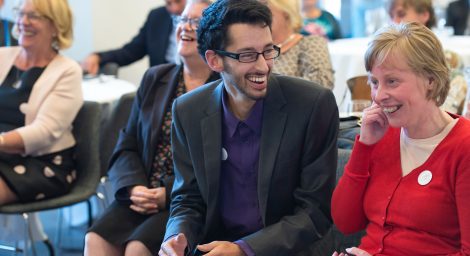 A young man seated at a busy event is smiling at a young woman sat next to him. They both look very happy and relaxed, as do the people we can see behind them.