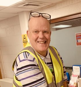 A man smiles joyfully at the camera. Only his head and shoulders are shown. He has glasses perched atop his head and is wearing a high vis vest. The background seems to be inside an office.